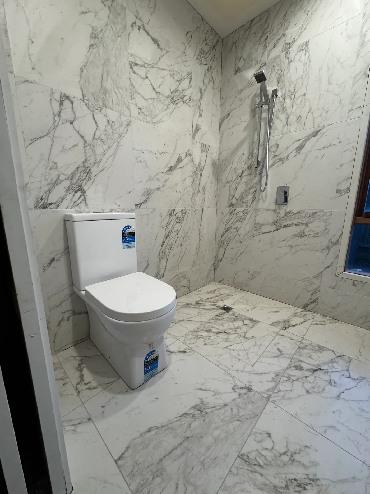 Newly fitted back-to-wall toilet suite and chrome rail shower in a bathroom tiled floor to ceiling in marble-look tiles