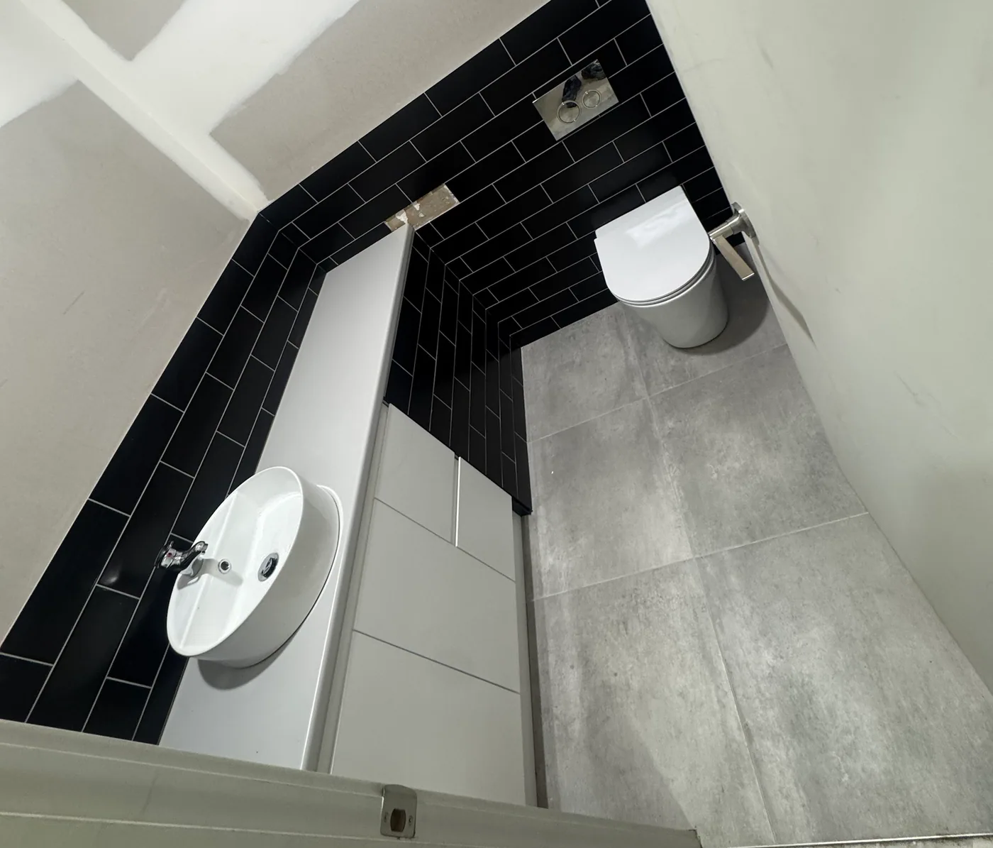 View down into a compact powder room with black subway wall tiles, a white wall-hung basin with chrome mixer and a wall-faced toilet