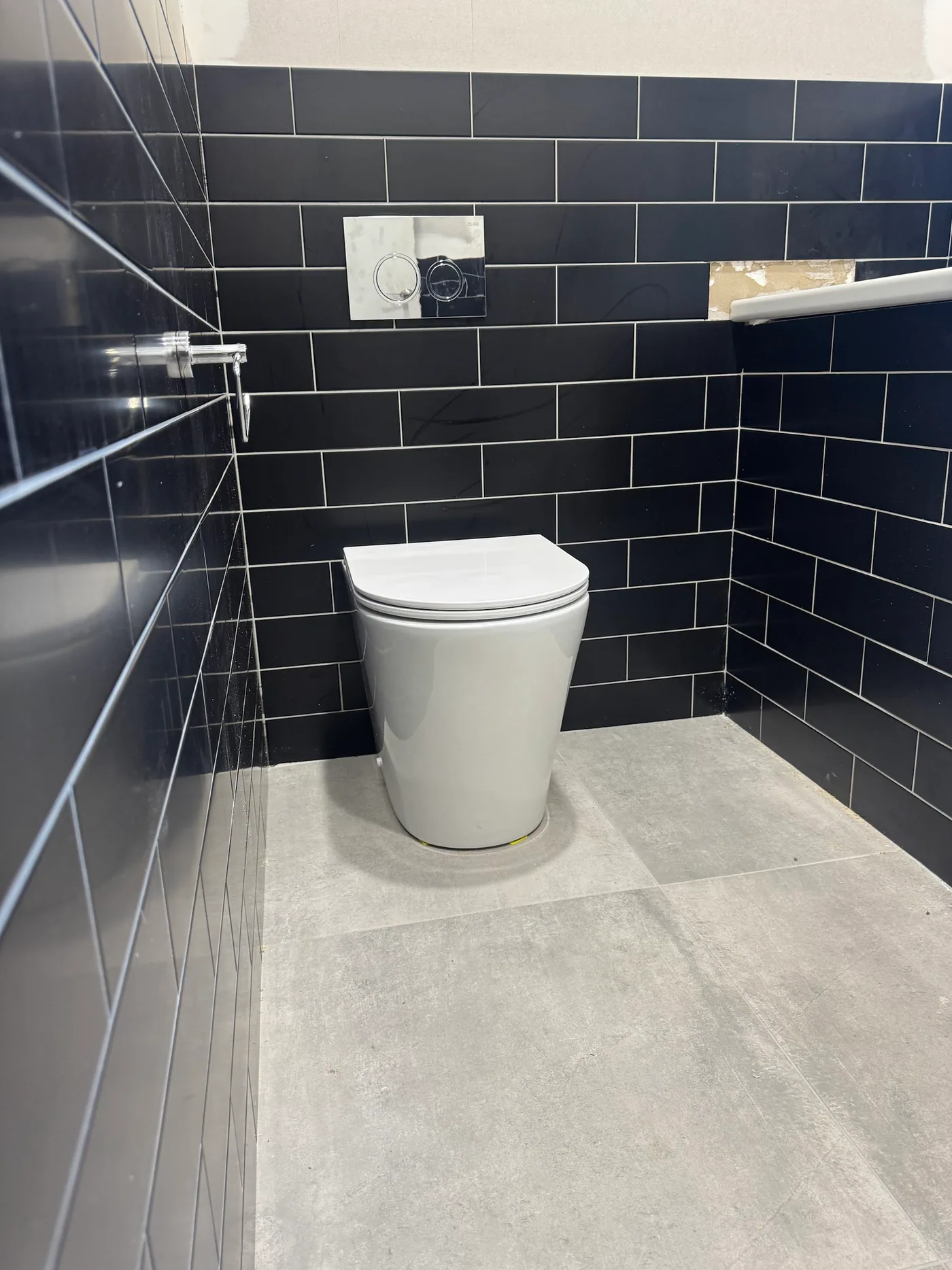 White wall-faced toilet pan with chrome dual flush plate on a black subway-tiled wall, grey concrete-look floor tiles