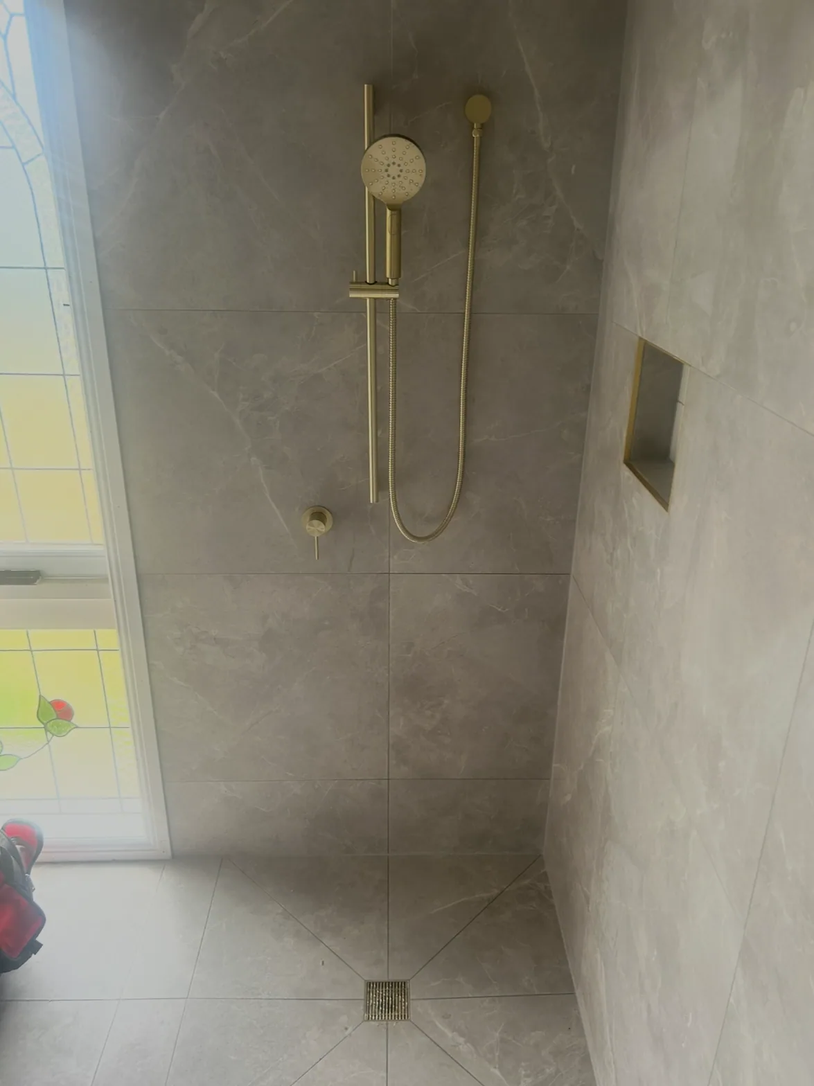 Shower with brushed brass rail shower, wall mixer and tiled niche, square floor grate centred in grey marble-look tiles